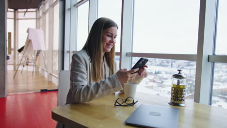 cheerful happy female sitting at the desk. smiling lady messaging to someone on the phone during tea break at work.
