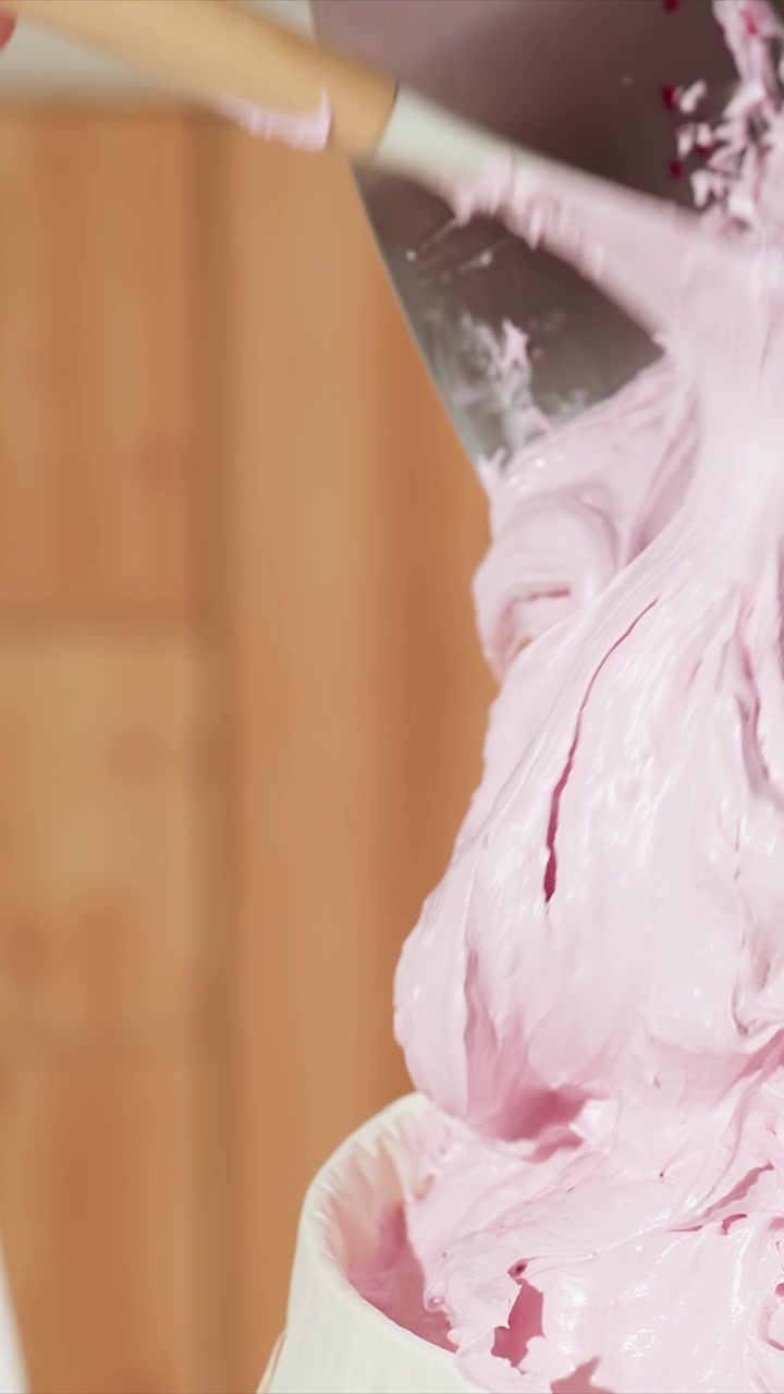 Primo piano di mani che usano una spatola per trasferire una crema rosa. La crema è densa e lucida, spostata agevolmente tra i recipienti. Ambiente cucina interno con pannelli in legno sullo sfondo.