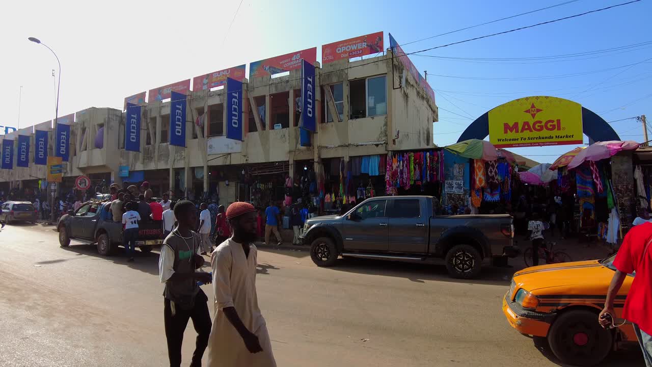Panning left across the entrance of Serekunda Market, showcasing shops and street activity with cars gathered outside of market as people walk along road
