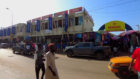 panning left across the entrance of serekunda market, showcasing shops and street activity with cars gathered outside of market as people walk along road