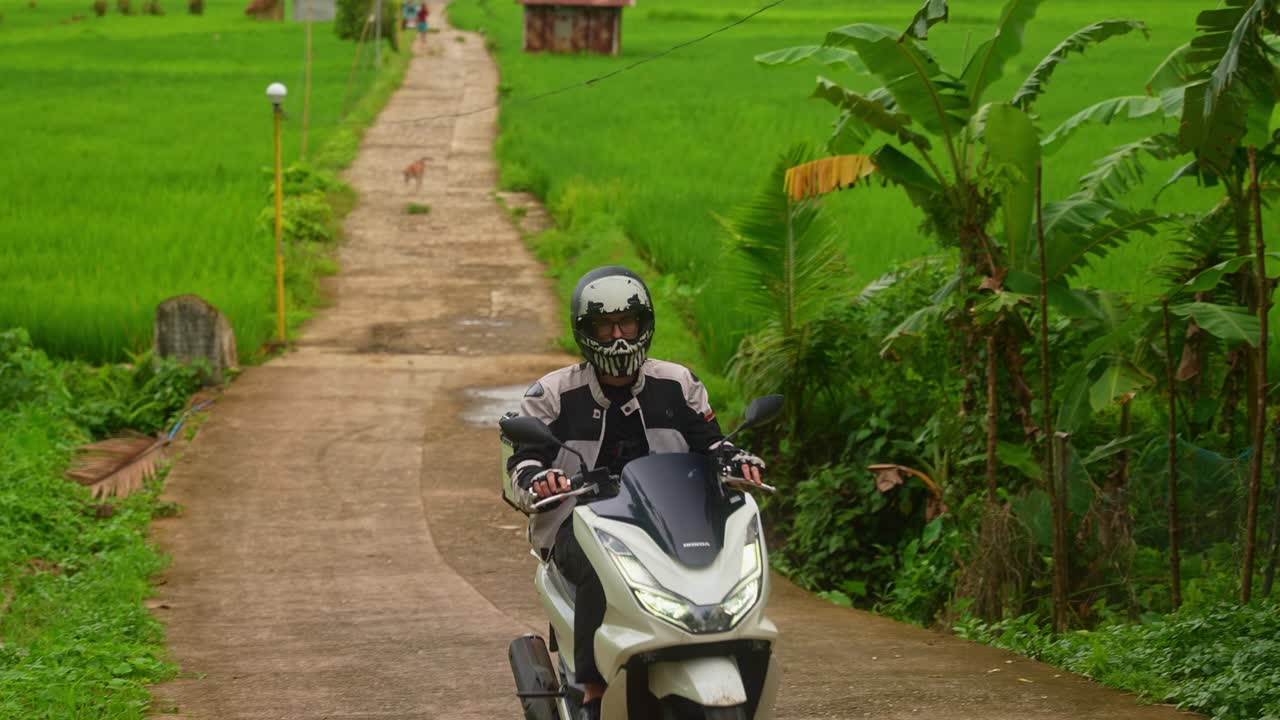 A focus shot of motorcycle rider back along Mahabang Tanaw in rice fields, briefly blinking lights toward the camera before riding off frame at the end in Quezon Province Philippines