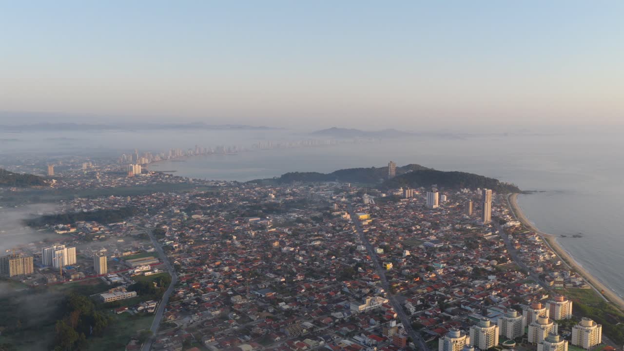 Ripresa aerea dall'alto con drone che si inclina verso il basso, inquadrando la disposizione della città costiera sullo sfondo dell'oceano e della nebbia mattutina