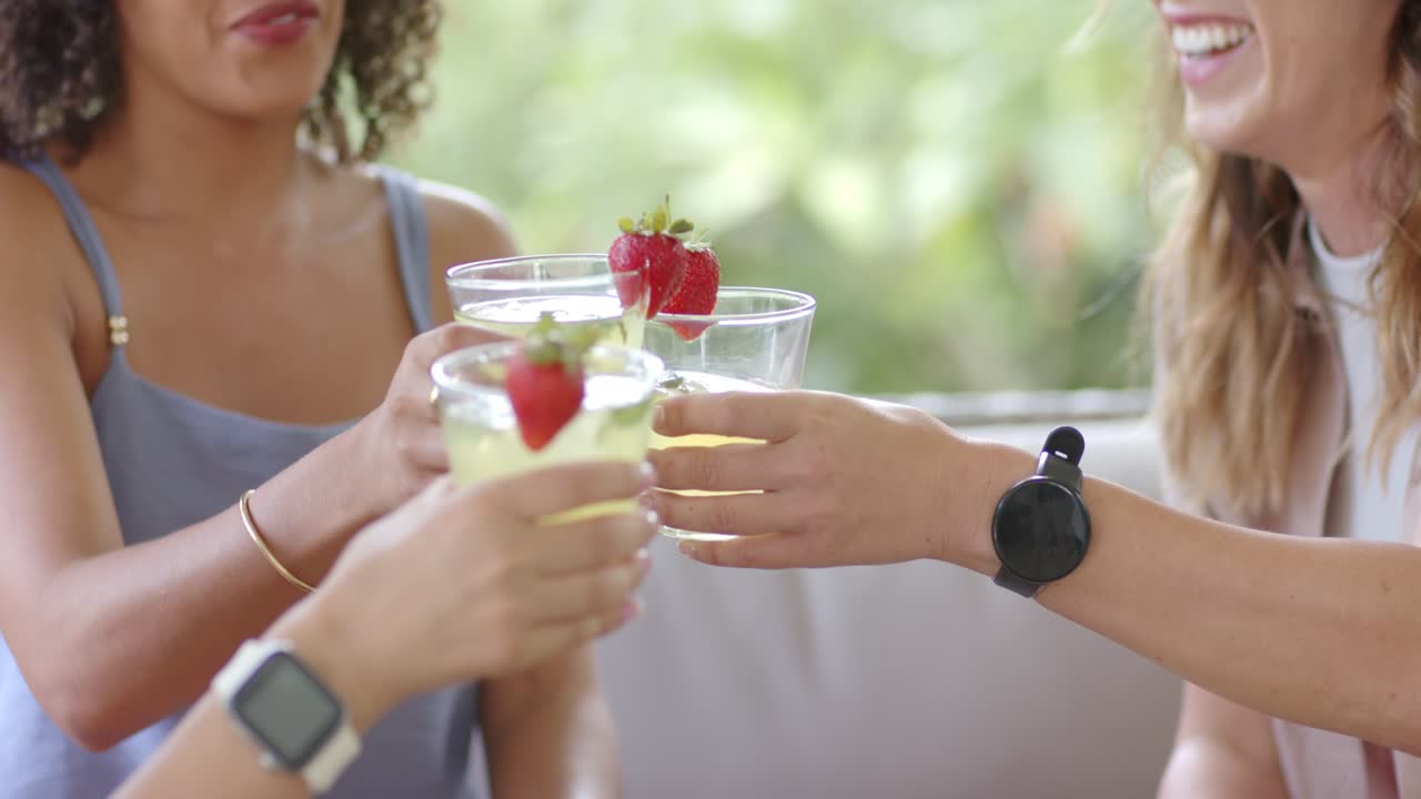 Diverse female friends on patio reacting to second arriving, raising strawberry drinks, toasting