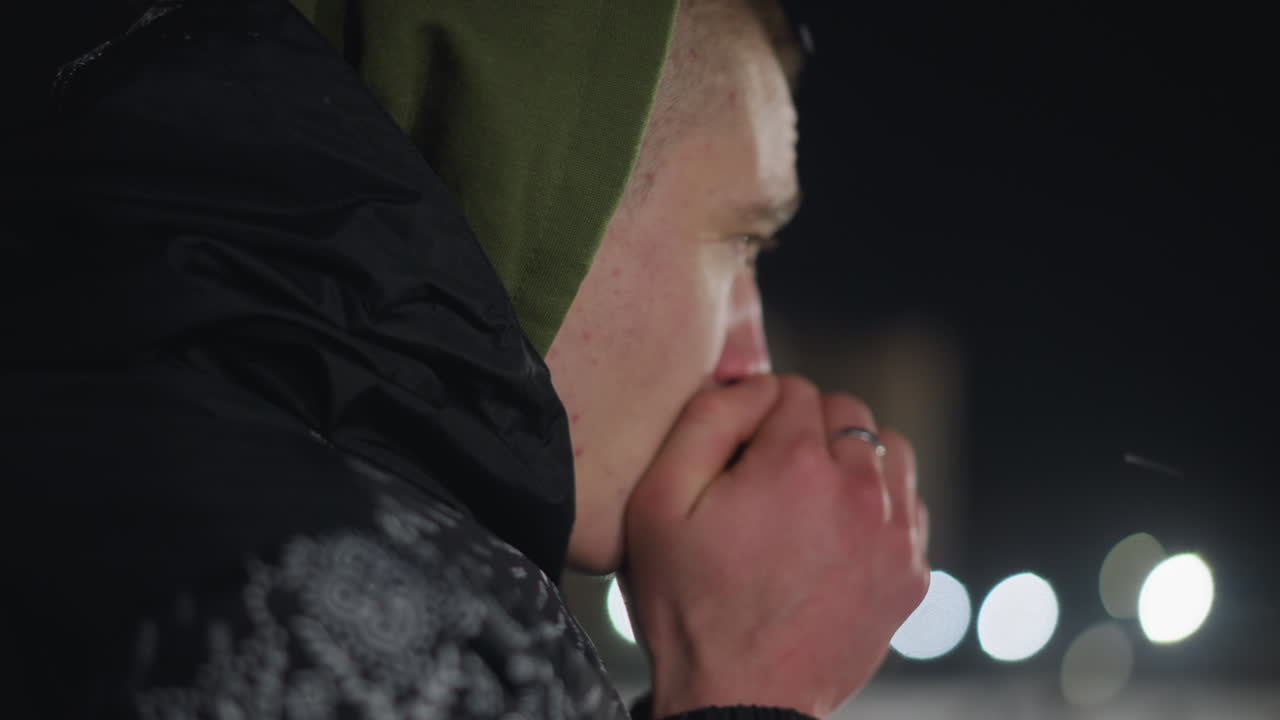 Close up side view of cold man in hooded jacket blowing warm air into hands as visible breath rises in freezing night with blurred glowing lights in background
