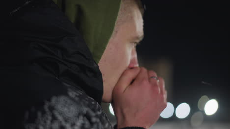 close up side view of cold man in hooded jacket blowing warm air into hands as visible breath rises in freezing night with blurred glowing lights in background