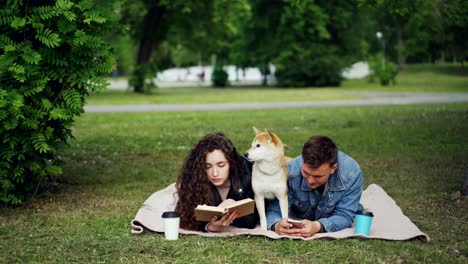 una joven familia pasando el fin de semana en el parque, una mujer leyendo un libro, un hombre usando un teléfono inteligente, un lindo perro descansando entre ellos. el tipo está mostrando la pantalla del teléfono a su esposa.