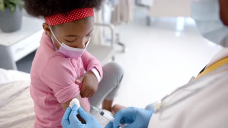 African-american-female-doctor-with-face-mask-making-injection-to-girl-in-hospital-room,-slow-motion