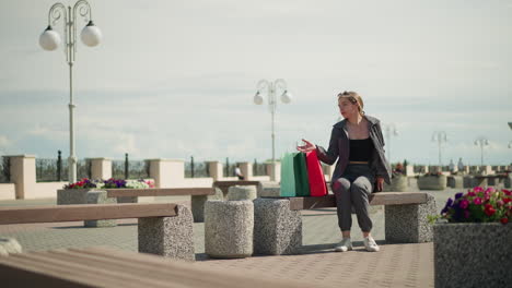 la señora se sienta en un banco al aire libre, deja caer sus bolsas de compras, ajusta la bolsa de compras roja, y se relaja con su mano en el banco, mira a la derecha, rodeada de bancos de parque y flores