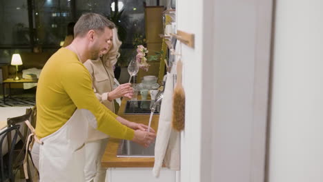 side view of a man washing the family dinner dishes at the sink in the kitchen while talking with a mature woman who is helping him