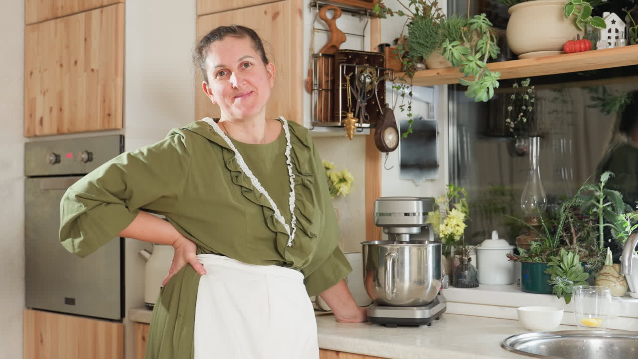 Chef stands in cozy kitchen beside running stand mixer with hand on waist and gentle smile, surrounded by flowers, herbs, and rustic decor, waiting patiently for batter to mix while enjoying moment