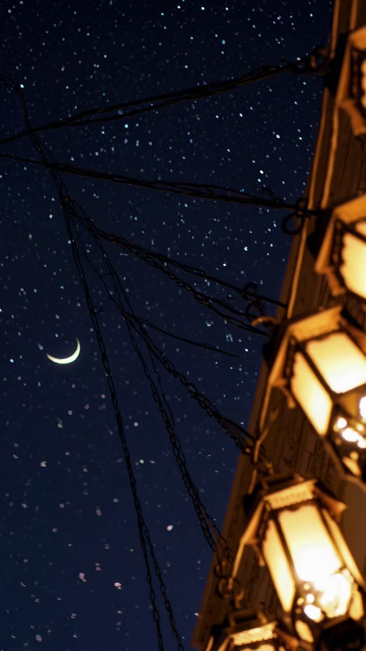 A dreamy night scene with hanging lanterns in the foreground and a crescent moon in the starry sky