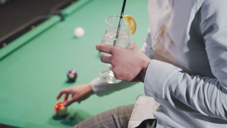 close-up of gentleman in white shirt and grey trousers holding lemon drink with black straw, sitting on green pool table with colorful billiard balls as he rolls a ball. soft lighting