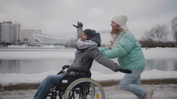 Happy woman running with her disabled friend in wheelchair and having fun together in the city in winter