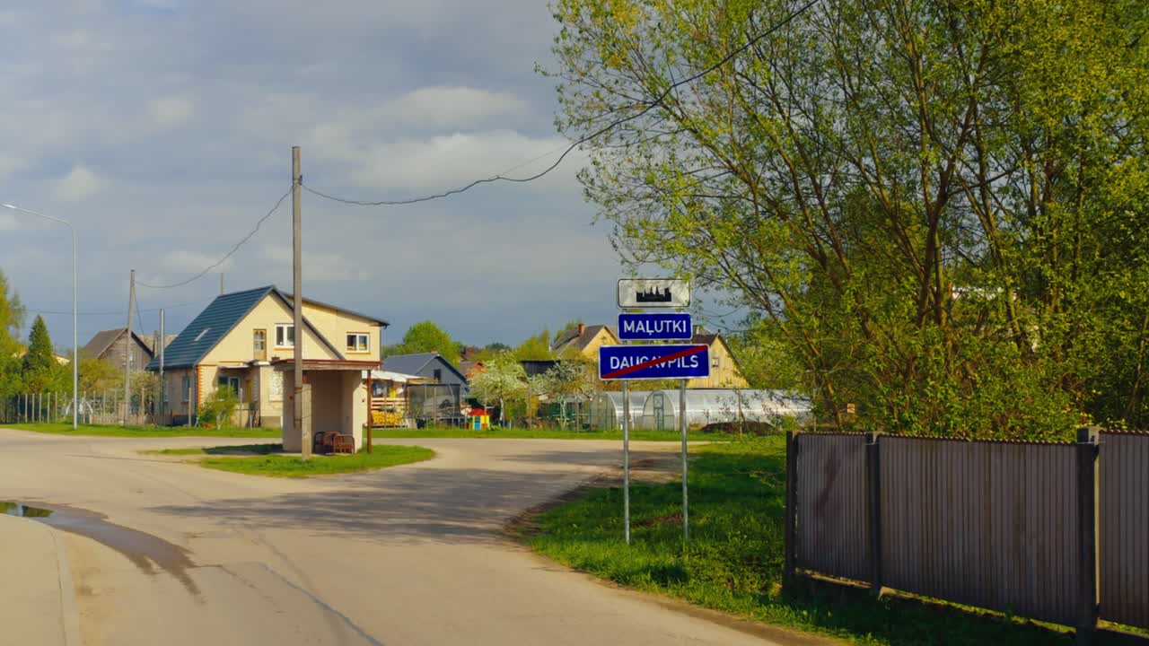 View of Malutki road sign and exit sign for Daugavpils beside village homes and bus shelter on a sunny afternoon. Malutki, Latvia (Latgale)