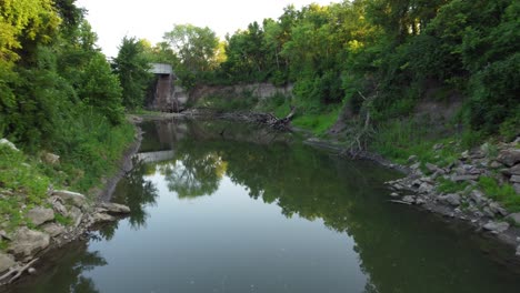 Drone-flying-around-creek-surrounded-by-trees-then-going-under-an-old-bridge