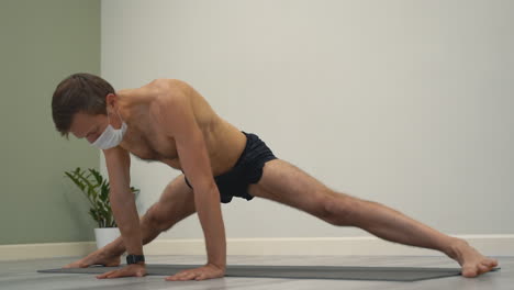 yoga at home. man practicing side split pose and kurmasana, wearing medical face mask, on white background with copy space. male athlete doing exercises indoors.