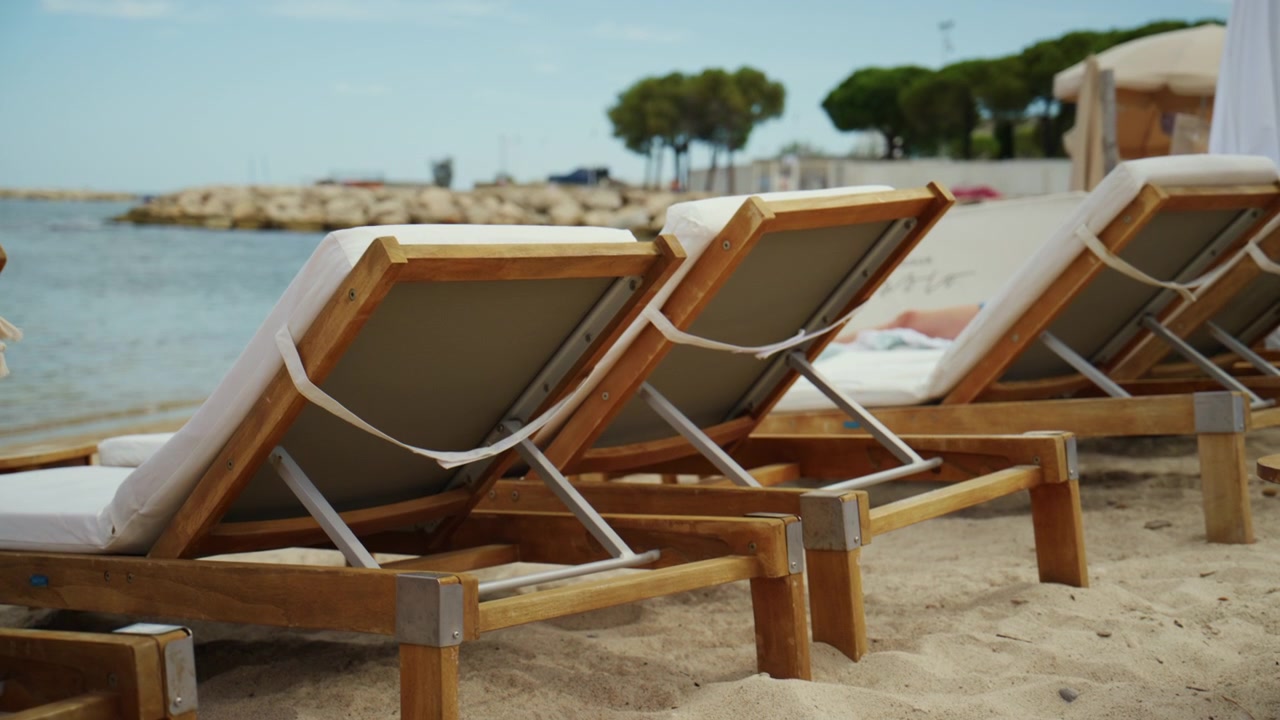 Chaises longues en bois avec des coussins blancs disposées sur le sable de la plage, offrant confort et tranquillité au bord de la mer.