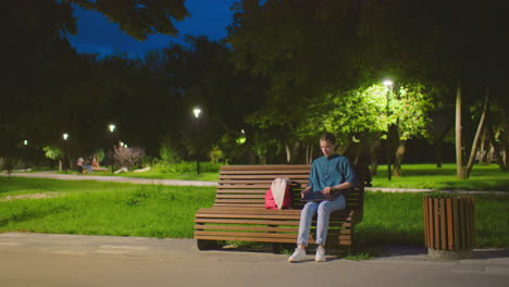 young lady sits outdoors on park bench with red bag nearby at dawn, opening laptop in tranquil setting, background includes illuminated trees, light poles, green lawn, and people strolling