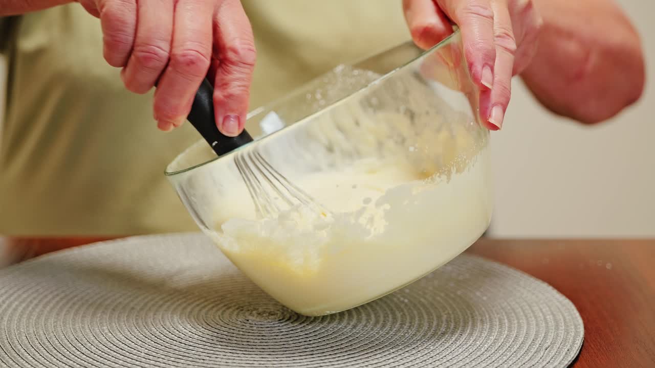 Slow motion hands whisk smooth cake batter in glass bowl on kitchen table