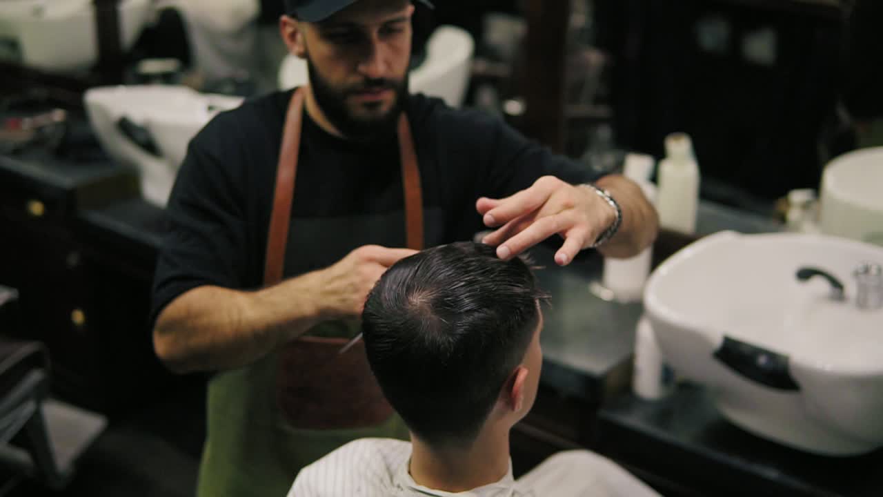 Bearded barber in apron combing hair of a male client sitting in the chair at barber shot during haircut. Stylish hairdresser