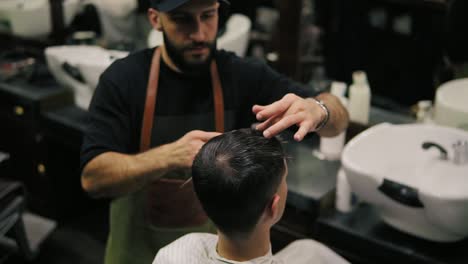 Bearded-barber-in-apron-combing-hair-of-a-male-client-sitting-in-the-chair-at-barber-shot-during-haircut.-Stylish-hairdresser