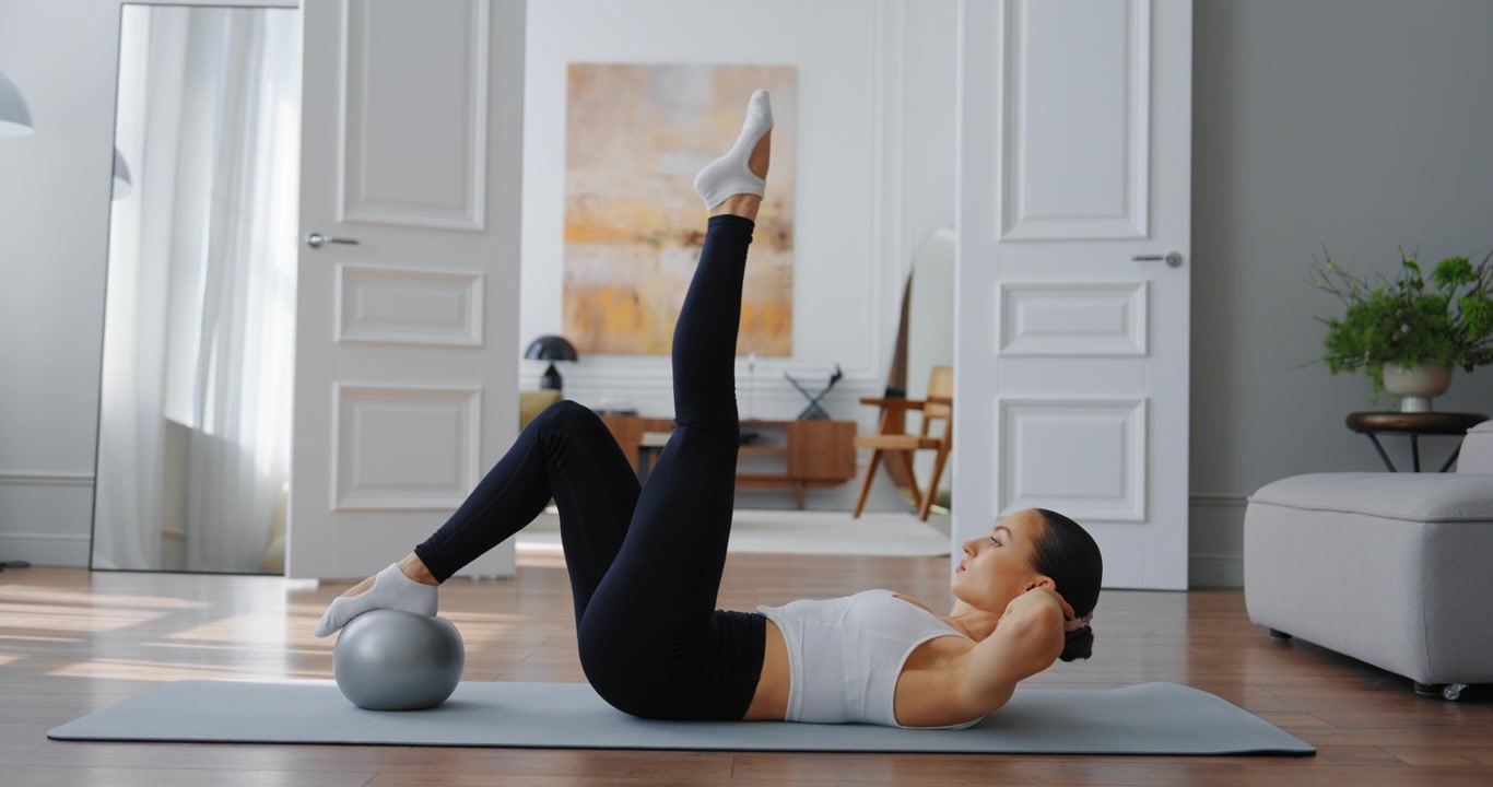 Giovane donna che esegue sollevamenti delle gambe con una palla da ginnastica sul tappetino da yoga a casa
