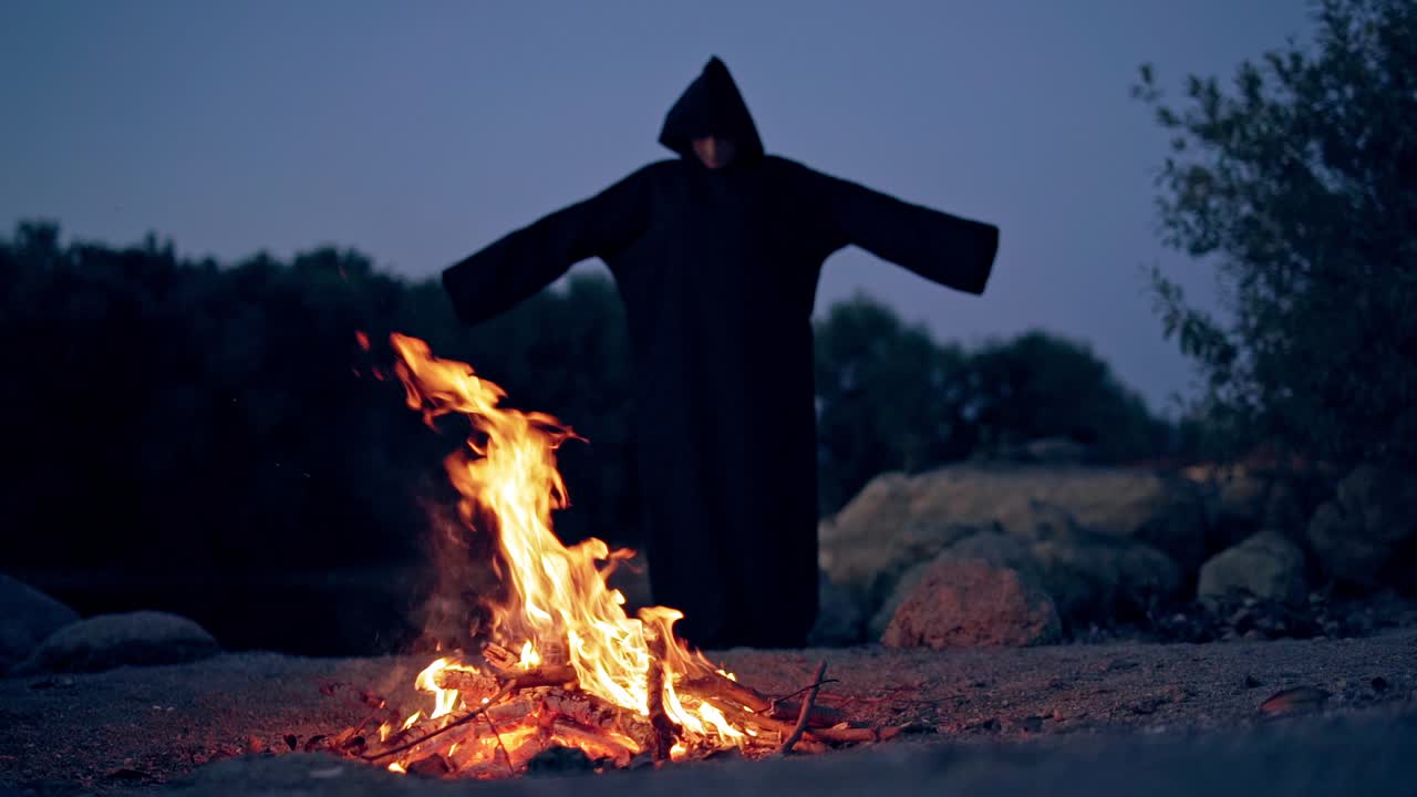 Scary view of a evil in black costume at the fire. Silhouette of death with stretched arms outdoors. Halloween evening.