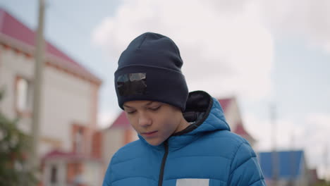 kid in black beanie and blue jacket strolling with head bowed thoughtfully, blurred background featuring buildings, light poles and trees