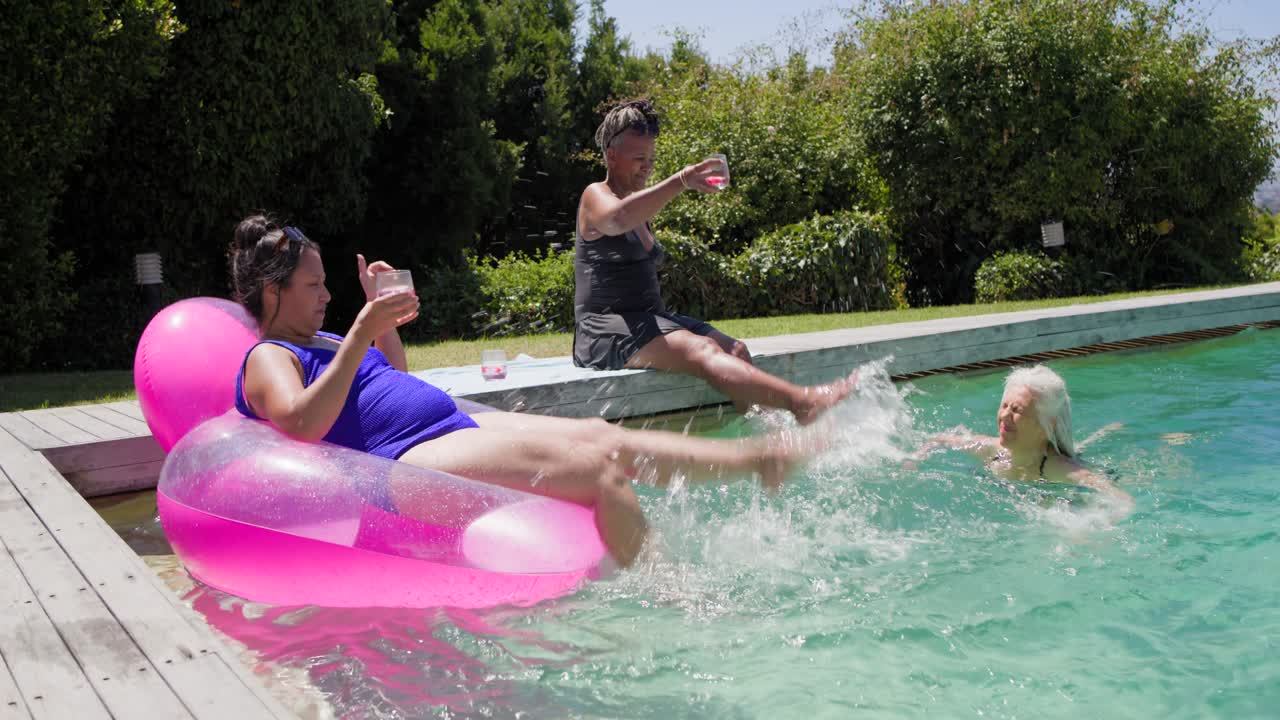 African American women in swimwear at pool reclining friend kicking float splashing senior for fun