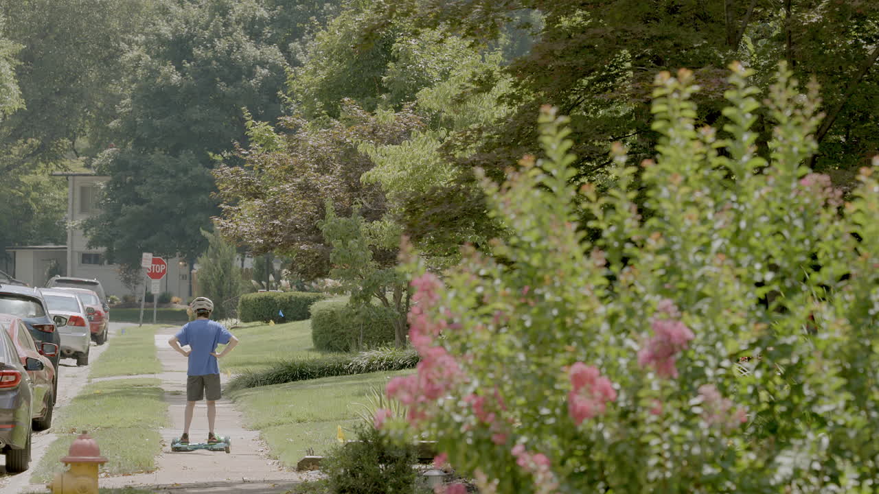 симпатичный молодой парень в велосипедном шлеме едет на ховерборде по тротуару в пригороде летним днем