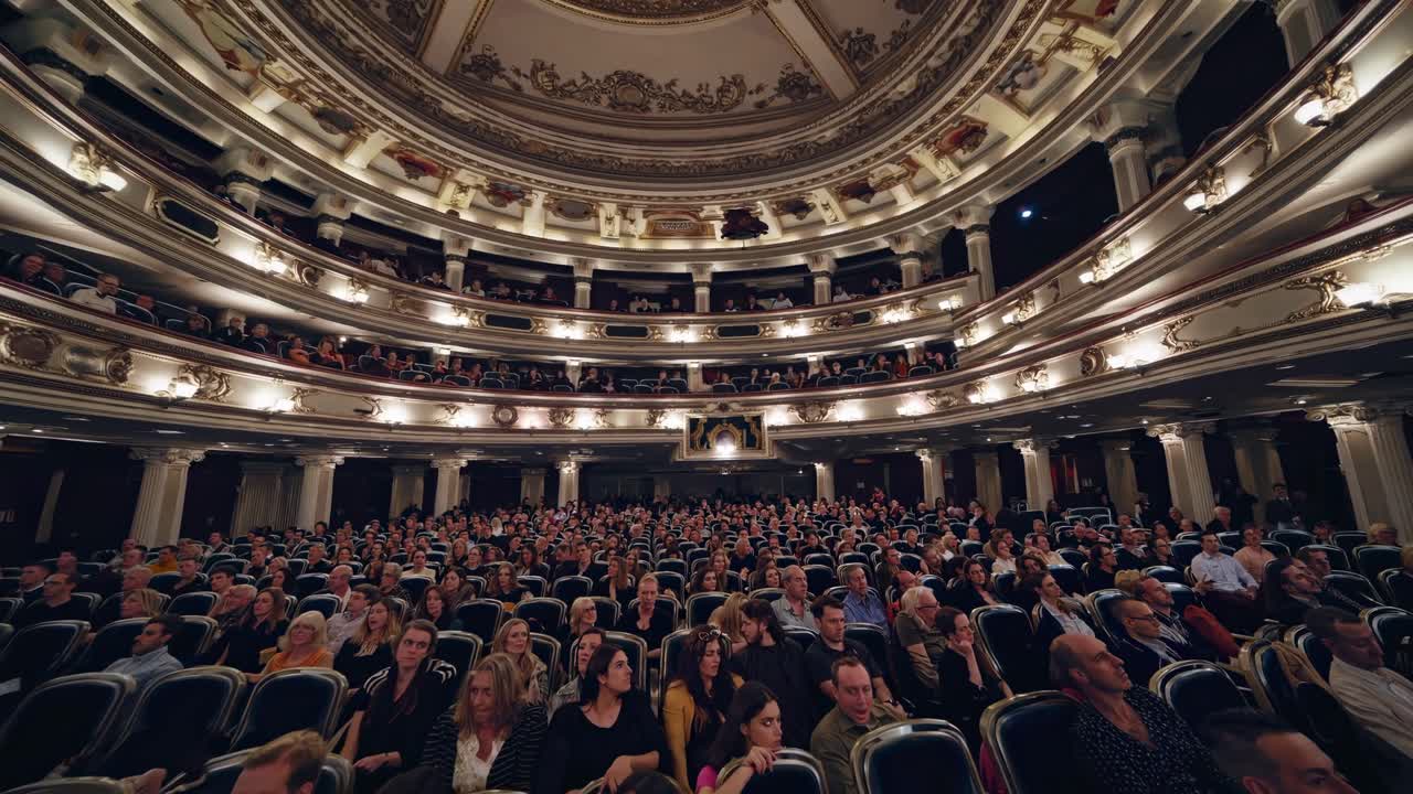 Inquadratura grandangolare che riprende un maestoso teatro pieno di pubblico, mettendo in risalto il soffitto ornato.
