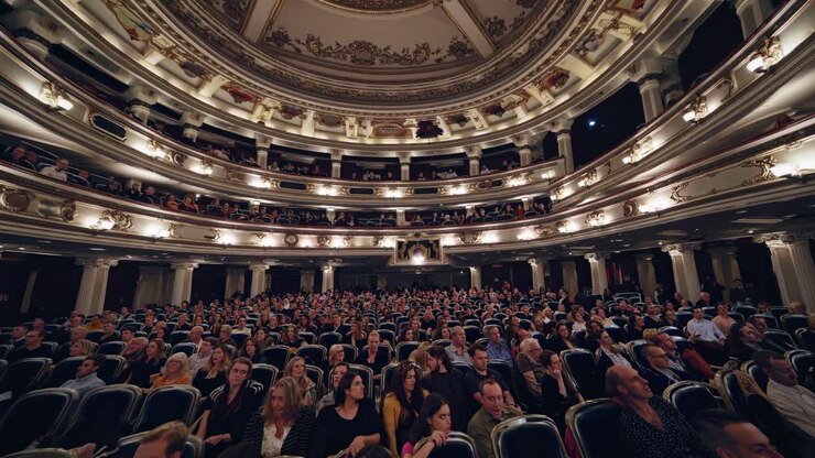Inquadratura grandangolare che riprende un maestoso teatro pieno di pubblico, mettendo in risalto il soffitto ornato.