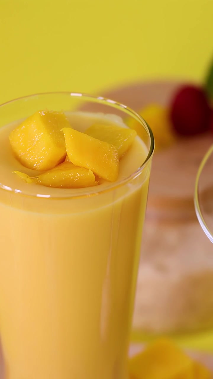 Primo piano di una mano che guarnisce un frullato al mango con cubetti di frutta su uno sfondo giallo brillante.