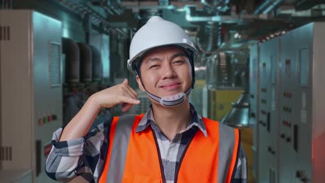 Close Up Of Asian Male Engineer With Safety Helmet Smiling To Camera And Making Call Me Gesture While Standing In Engine Control Room, Work Of Electrical Generators