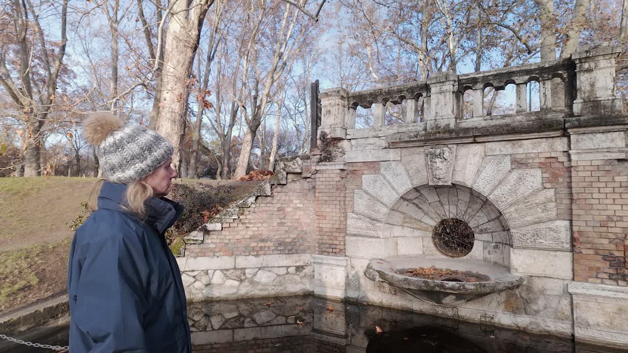 una mujer con sombrero admira una vieja fuente en el parque de budapest en el frío de otoño.