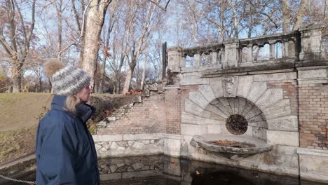 woman in hat admires old fountain in budapest park in autumn chill