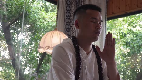 a man meditates indoors with eyes closed, wearing white robes and wooden beads, surrounded by greenery visible through large windows