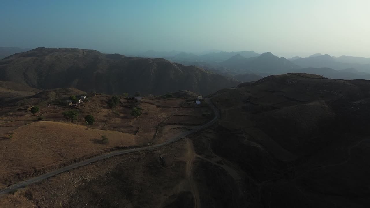 Avvicinamento aereo su colline aspre vicino a Udaipur, India, rivelando strade sterrate tortuose, creste montuose aride e orizzonti stratificati in una calda luce radente.