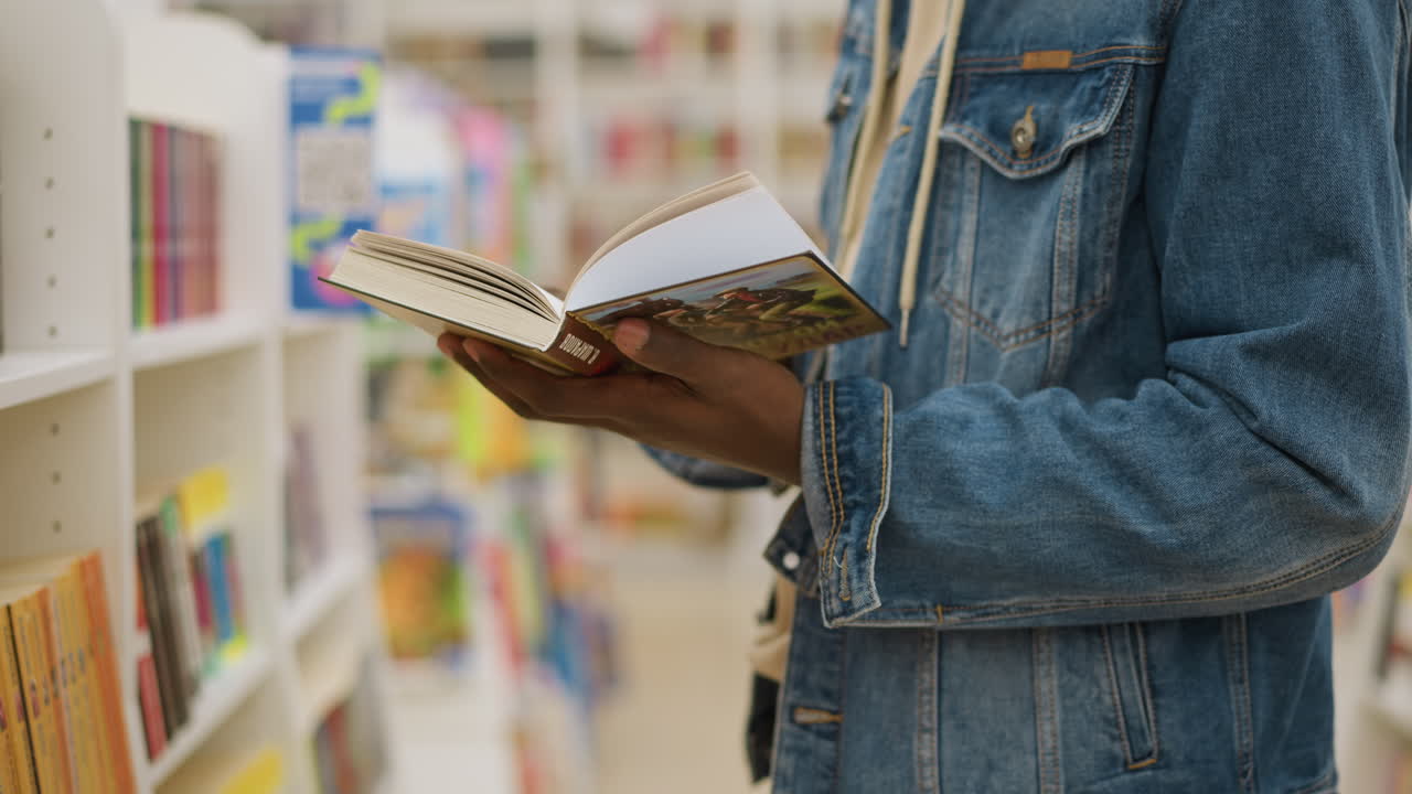 Man in denim jacket holding open book in bookstore, browsing pages while standing in front of shelves filled with colourful books, capturing the joy of reading in a vibrant, educational setting