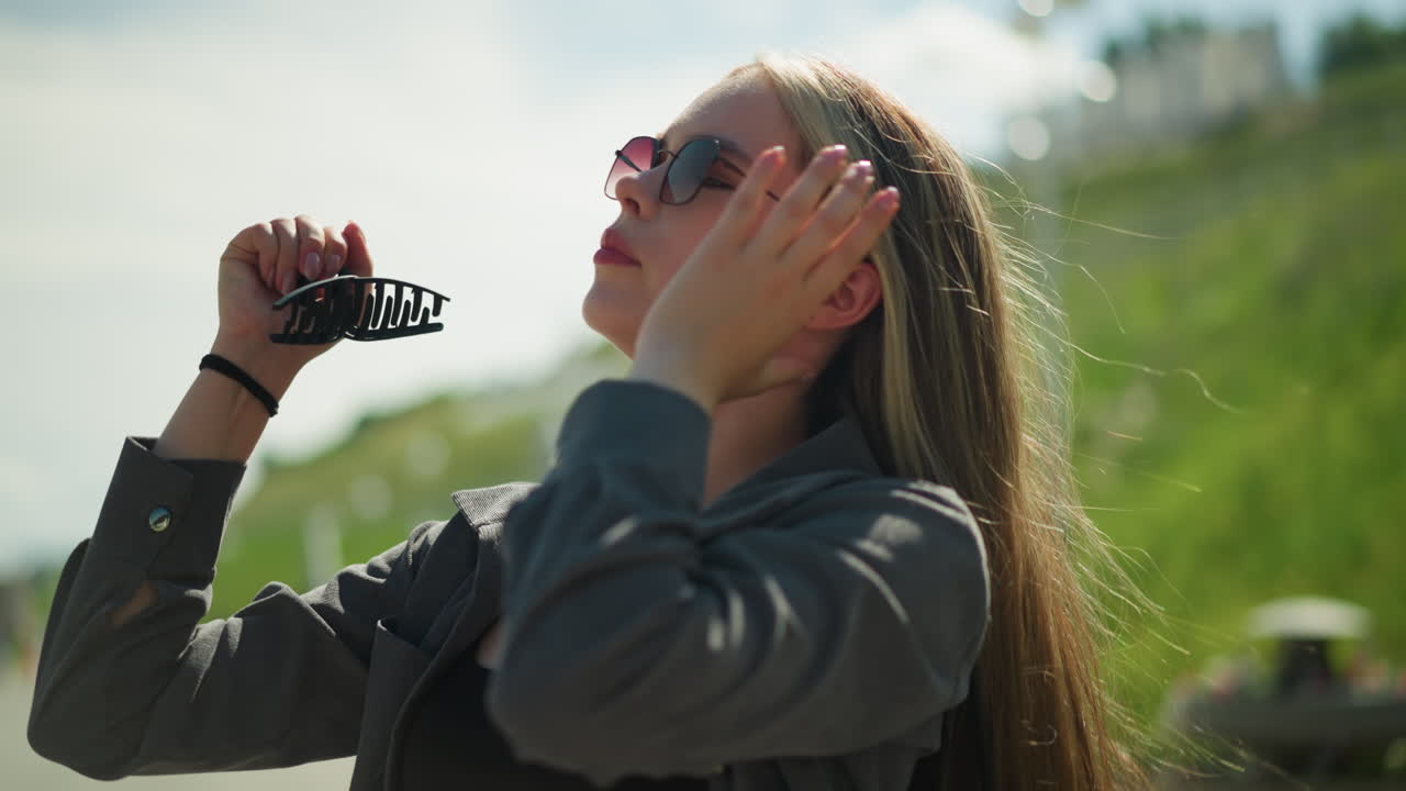 Lady in black top under grey jacket, wearing sunglasses, trying to clip her hair with a large hair clip, her focus is on arranging her hair as she stands on a sunny day, with blur view of hill
