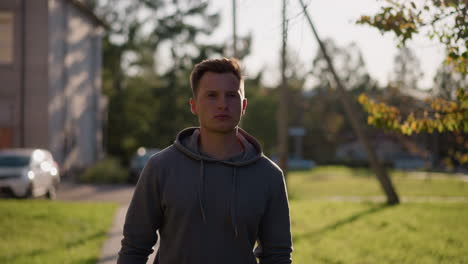 young man in dark gray hoodie walking outdoors in sunny park with serious expression, surrounded by green grass, trees, distant building in soft focus background, autumn setting, relaxed mood