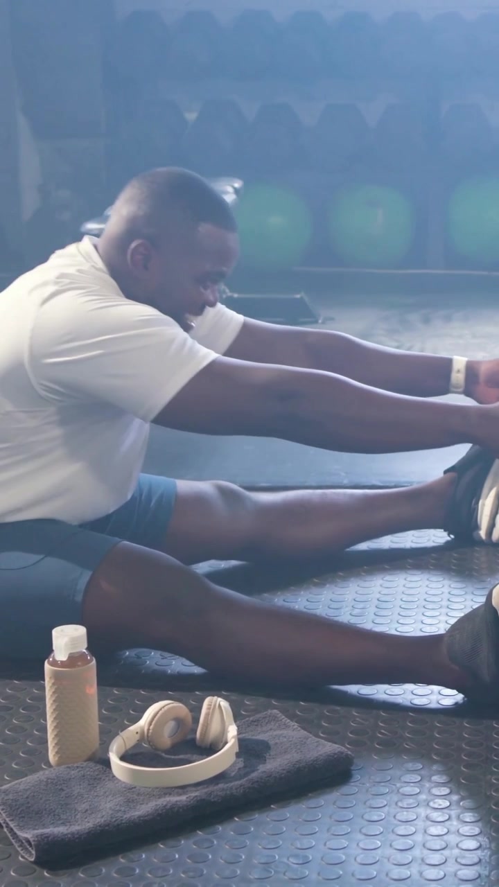 Vertical video: Stretching man after workout, reaching on gym floor, with bottle, balls, bench