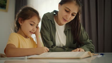 A-little-brunette-girl-with-a-braid-hairstyle-in-a-yellow-T-shirt-does-her-homework-and-writes-in-her-notebook-together-with-her-mother,-a-brunette-girl,-in-a-Green-jacket-and-a-white-T-shirt-on-a-white-table-in-a-modern-children's-room