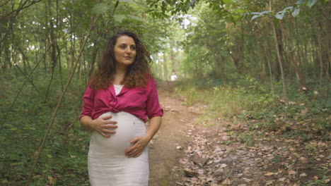 pregnant woman expectant mother walking alone in forest, attractive caucasian casual dressed mom stroking caressing touching her belly with hands smiling, childbearing tenderness, medium portrait shot