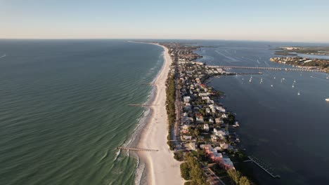 Rising-drone-clip-over-thin-peninsula-with-buildings-and-white-sandy-beach,-surrounded-by-open-sea,-connected-by-road-bridge