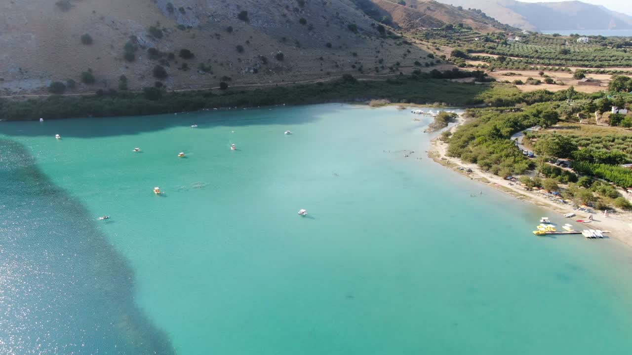 Drone view in Greece flying over a light and dark blue lake with small boats and surrounded by green mountain on a sunny day in Crete