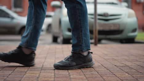 side view of guy wearing blue jeans walking along tiled urban walkway beside parked cars and red brick buildings, focus on shoe motion and footstep pattern during cold city scene