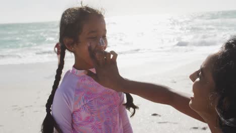 Glückliche-Hispanische-Mutter,-Die-Am-Strand-Sonnencreme-Auf-Das-Gesicht-Ihrer-Tochter-Aufträgt