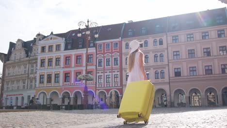 rear view of elegant woman walking in old city, pulling yellow suitcase. female tourist with bright yellow suitcase on old cobblestone. vacation in a european city