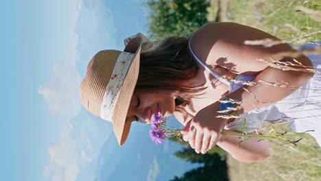 Vertical-shot-of-pretty-girl-smelling-blooming-flower-in-mountains-during-summer-day
